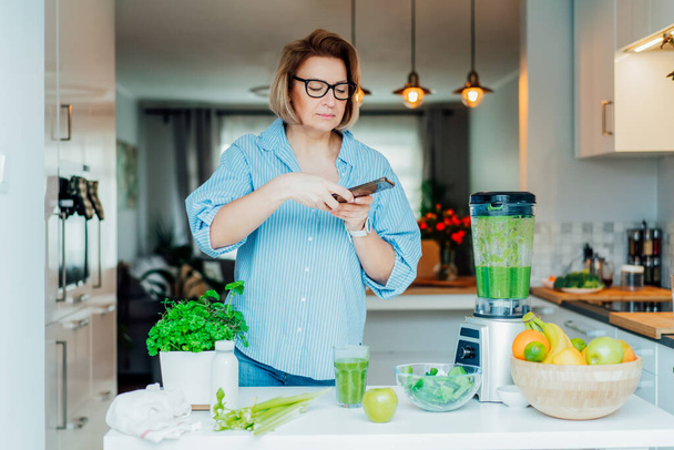 Blogger creating content for social media. Woman making photo, video with just made glass of detox green smoothie and its ingredients on the kitchen table. Healthy dieting, cooking, weight loss blog - Photo, image