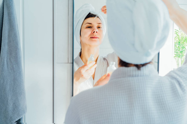Mixed raced woman in bath spa towel applying rejuvenation moisturizing serum on her face looking at the mirror at home after hot bath and shower. Beauty treatment, skincare, home Beauty self-care. - Fotografie, Obrázek