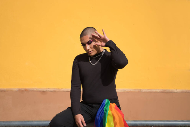 Niebinarna osoba, młody Amerykanin z Ameryki Południowej, mocno przerobiony, siedzący na balustradzie z flagą dumy gejowskiej trzymaną w kieszeni spodni. Królowa koncepcji, lgbtq +, duma, queer. - Zdjęcie, obraz