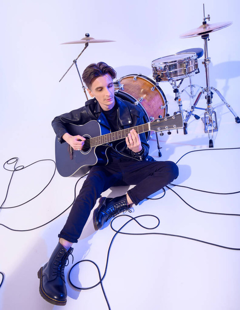 Молодой парень, играющий на акустической гитаре, сидит на полу, опираясь на барабан - Фото, изображение