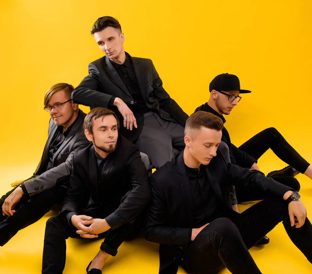Grupo de cinco chicos jóvenes en trajes negros sobre un fondo amarillo  - Foto, Imagen