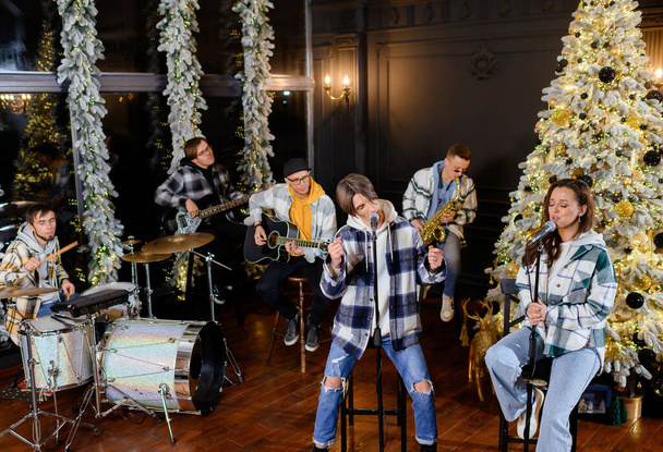 Genç rock grubu Noel ağacının arka planında şarkı söyler ve çalar.  - Fotoğraf, Görsel