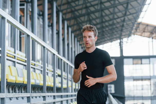Running on the bleachers. Sportive young guy in black shirt and pants outdoors at daytime. - Foto, Bild