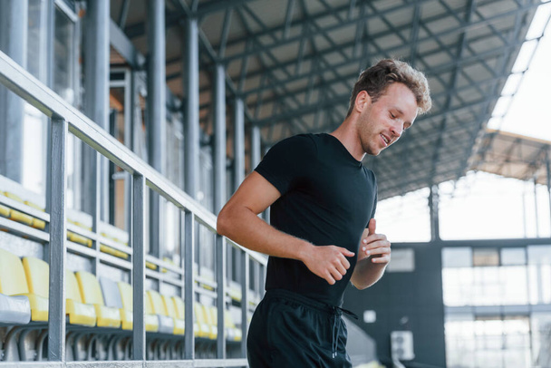 Running on the bleachers. Sportive young guy in black shirt and pants outdoors at daytime. - Foto, Imagen