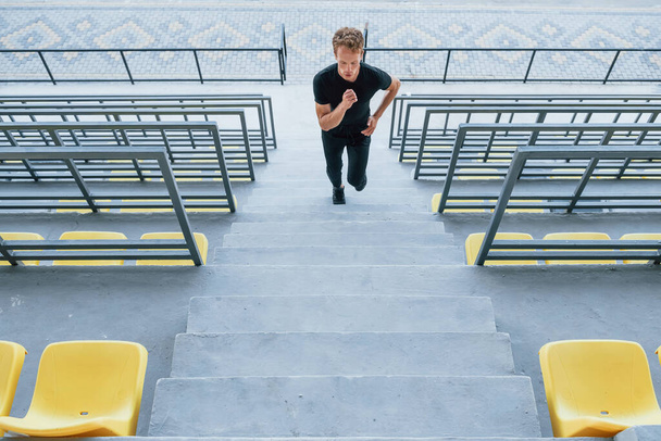 Running on the bleachers. Sportive young guy in black shirt and pants outdoors at daytime. - 写真・画像