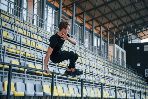 Jumping and doing parkour on the bleachers. Sportive young guy in black shirt and pants outdoors at daytime. - 写真・画像