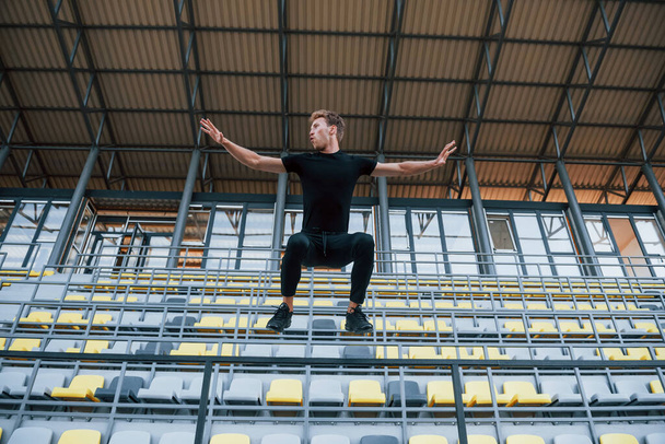 Jumping and doing parkour on the bleachers. Sportive young guy in black shirt and pants outdoors at daytime. - Zdjęcie, obraz
