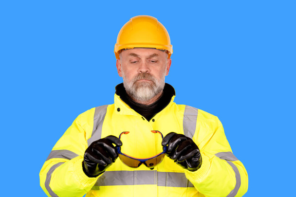 Sarı kasklı bir inşaat işçisi, sarı bir yüksek çözünürlüklü ceket, mavi arka planda koyu renkli gözlük takmaya hazır. Vizyon güvenliği poster konseptini korumak için inşaat alanında doğru göz korumasını takın - Fotoğraf, Görsel