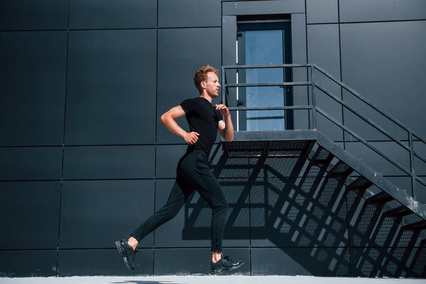 Running near black building. Sportive young guy in black shirt and pants outdoors at daytime. - Fotoğraf, Görsel