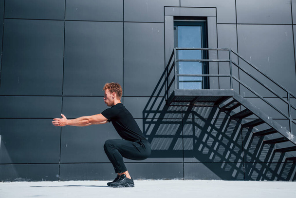 Exercising near building with black wall. Sportive young guy in black shirt and pants outdoors at daytime. - Foto, imagen
