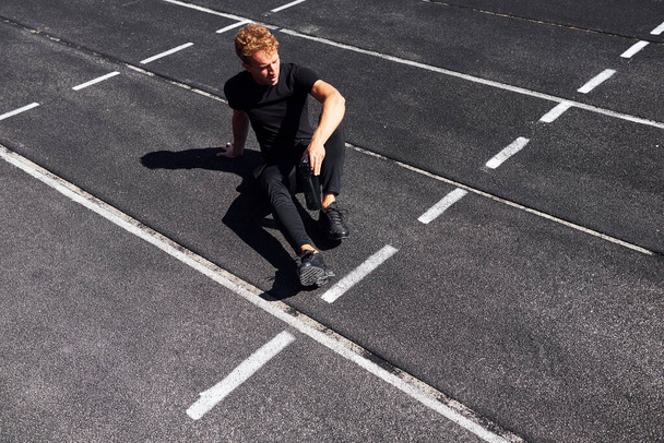 Tired runner sits on track and taking a break. Sportive young guy in black shirt and pants outdoors at daytime. - Foto, immagini
