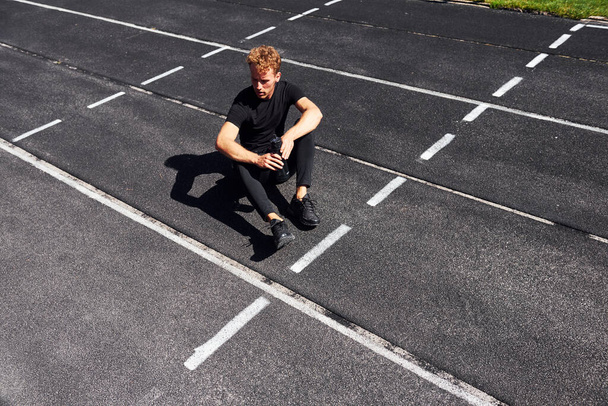 Tired runner sits on track and taking a break. Sportive young guy in black shirt and pants outdoors at daytime. - Fotoğraf, Görsel