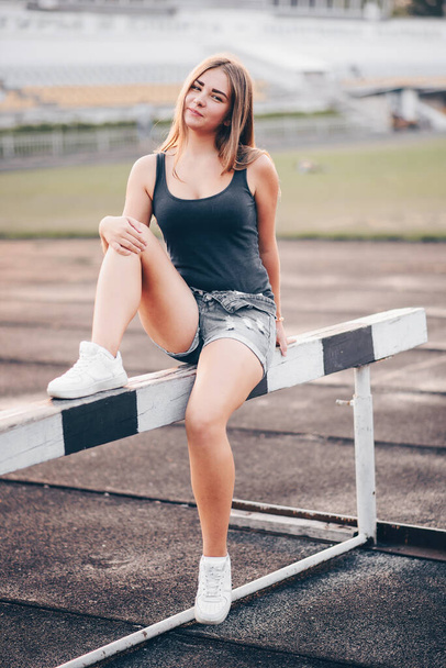Athlete sits on a Hurdle in white sneakers in a stadium with a smile on her face. A girl with long hair poses at the stadium at sunset. - Foto, Imagen