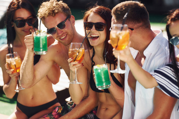 Fresh cocktails in hands. Group of young happy people have fun in swimming pool at daytime. - Foto, afbeelding