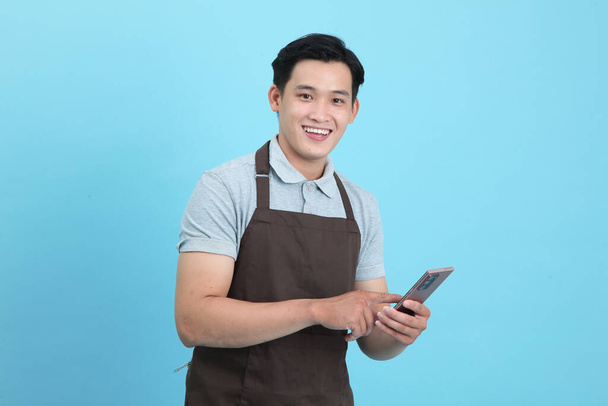 Asian young handsome man in waiter uniform, welcome guest and take order, isolated on blue background - Foto, Bild