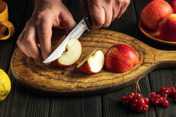 Ο σεφ κόβει μήλα σε ένα ξύλινο ξύλο κοπής με ένα μαχαίρι για να φτιάξει κομπόστα ή χυμό φρούτων. Μήλο διατροφή για ένα σύνολο βιταμινών. - Φωτογραφία, εικόνα