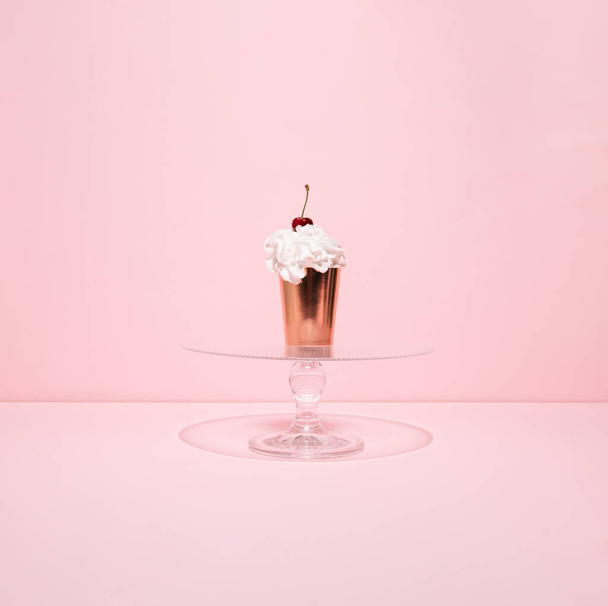 Golden paper cup filled with whipped cream and cherry on the top on glass cake stand against pink background. Summer cupcake. Minimal concept. - Photo, Image