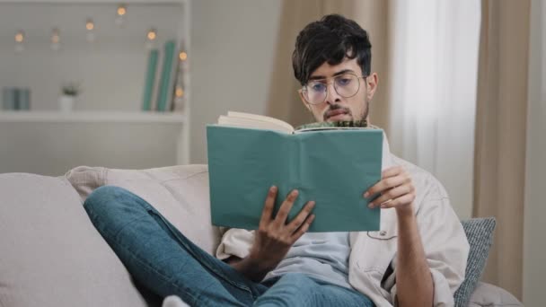 Молодой парень студент умный концентрированный арабский испаноязычный мужчина мужчина араб носит очки, лежащие на диване дома, читая книгу наслаждаясь литературой с учебником в помещении, хобби концепции обучения - Кадры, видео