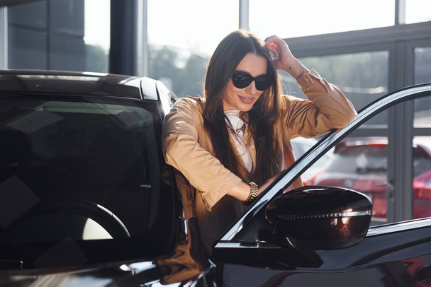 Standing near opened window. Fashionable beautiful young woman and her modern automobile. - Foto, afbeelding