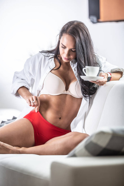 Woman in red menstruation panties and white bra is happy with the product holding a coffee on a sofa at home. - Foto, immagini