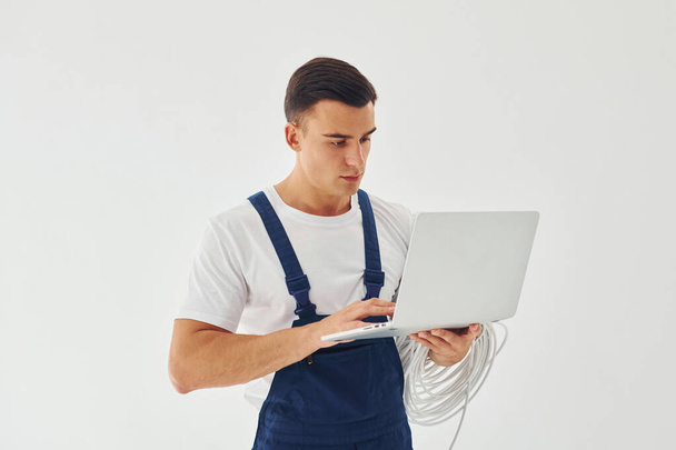 Uses laptop. Male worker in blue uniform standing inside of studio against white background. - 写真・画像