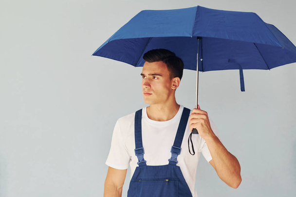 Holds umbrella by hand. Male worker in blue uniform standing inside of studio against white background. - Фото, изображение