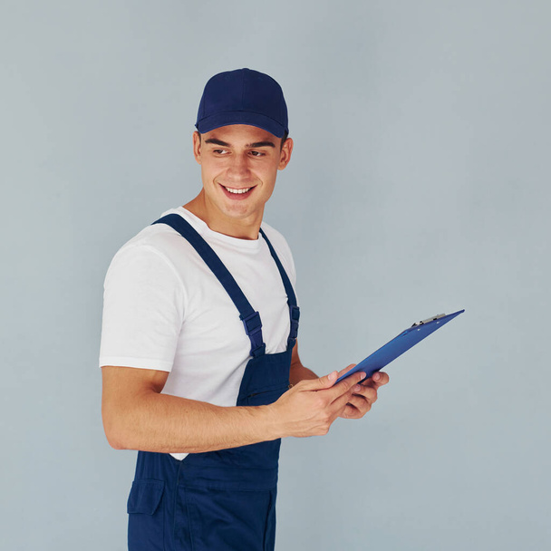 In cap and with notepad. Male worker in blue uniform standing inside of studio against white background. - Fotografie, Obrázek