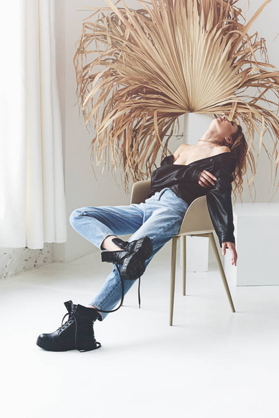Fashionable woman model dressed in jeans, leather jacket sitting on chair, palm leaf on a background - 写真・画像