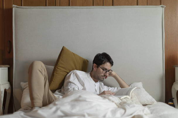 Człowiek leży w łóżku czytając książkę, odżywiając swój intelekt i ciesząc się przyjemnym wypoczynkiem w domu. - Zdjęcie, obraz