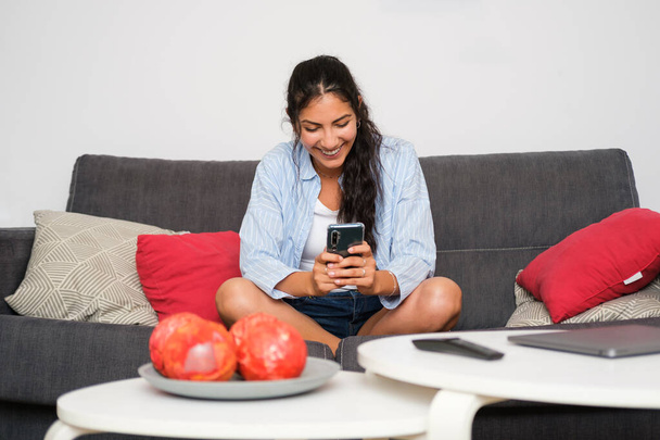 Relaxed casual latin woman texting or doing video call on her smartphone sitting on her couch at home. - Фото, изображение