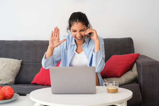 Young excited latin casual woman on mobile phone call using laptop at home. Expressive girl gesturing. - Fotoğraf, Görsel