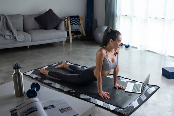 Sportliche junge Asiatin plankt vor Laptop, Wohnzimmereinrichtung, Seitenansicht, Kopierraum. Gesunde junge Frau in Sportbekleidung, die zu Hause Sport treibt und Online-Fitnesskurse anschaut - Foto, Bild