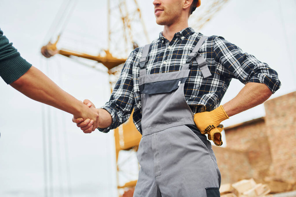 Making handshake. Two construction workers in uniform and safety equipment have job on building together. - Zdjęcie, obraz