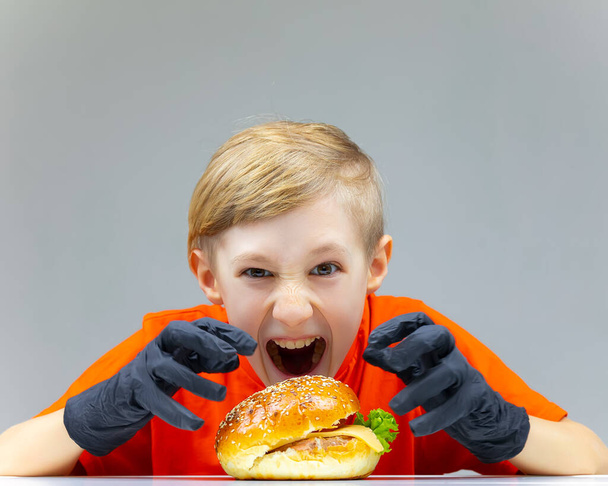 un chico con una hamburguesa con queso hace caras mostrando que tiene hambre - Foto, Imagen