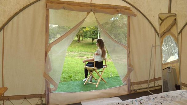 Вид из современной палатки на путешествующую женщину, сидящую на раскладном стульчике за пределами современной палатки, отдых и наслаждение природой в парке диких сафари. Летние приключения, отдых на природе, экотуризм, цифровой детокс. - Фото, изображение