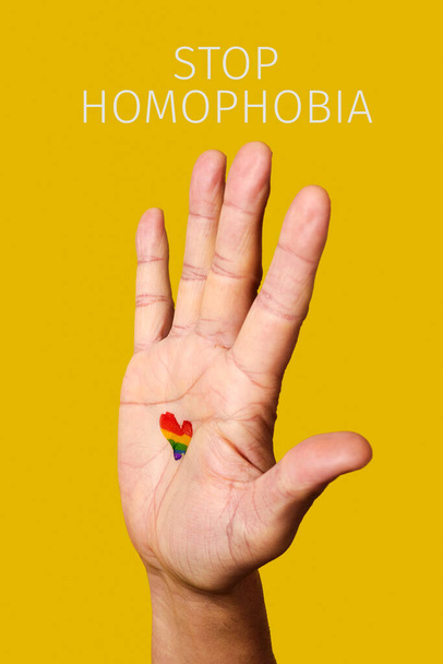 tekst zatrzymuje homofobię i podniesioną rękę mężczyzny, z tęczową flagą w kształcie serca namalowaną w dłoni, na żółtym tle - Zdjęcie, obraz