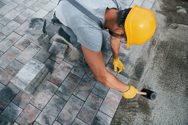 Uses a rubber hammer. Male worker in yellow colored uniform have job with pavement. - Foto, imagen