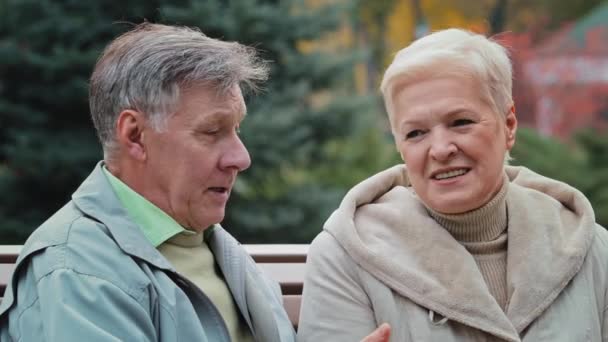 Elderly caucasian married couple lovely grandparents family sitting on bench in autumn park enjoying carefree conversation spend time together outdoors retired husband man talk with beloved woman wife - Footage, Video