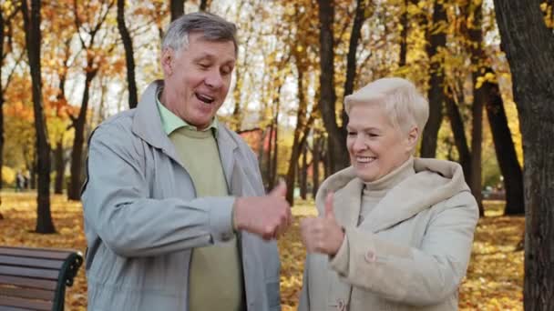 Portré boldog idős kaukázusi család felesége férj idősebb nagyszülők állni szabadban ősszel park mosolygós öreg pár idősebb férfi nő mutat hüvelykujj fel kéz gesztus jóváhagyás ajánlás - Felvétel, videó