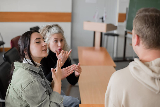 Two girls and a guy are talking in sign language. Three deaf students chatting in a university classroom. - Фото, зображення