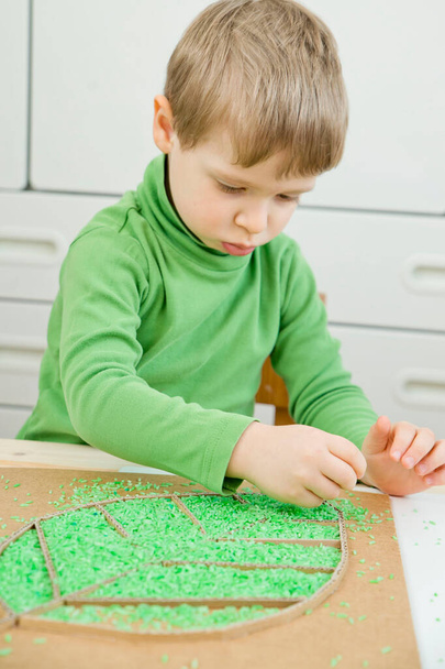 El chico juega con arroz de color verde. Implementar para que los niños desarrollen habilidades motoras finas a través del juego. Juguete de bricolaje para aprender la mano - coordinación ocular. - Foto, Imagen