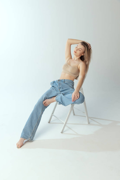 Portrait of tender young girl sitting on chair, posing in beige underwear and big jeans isolated over grey studio background. Self-love - Foto, immagini