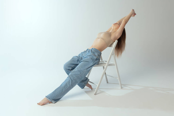 Tender beautiful young girl posing in underwear and jeans on chair isolated over grey studio background. Self-love - Фото, изображение