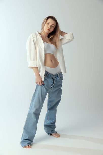 Full-length portrait of young slim girl wearing shirt, underwear and jeans isolated over grey studio background. Youth beauty - Foto, imagen