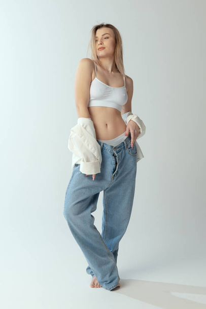Full-length portrait of young slim girl wearing shirt, underwear and jeans isolated over grey studio background - Fotó, kép