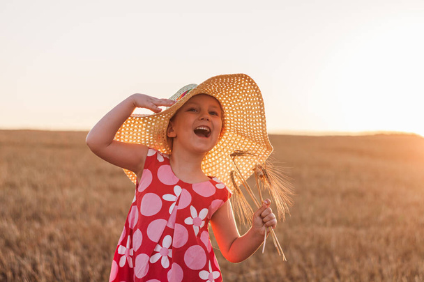 Egy szalmakalapos kislány búzapecsenyébe öltözve. Mosolygó gyerek napszemüvegben a naplemente táján. Családi gazdálkodás mezőgazdasági környezet ökológia koncepció. Cottagecore stílusú esztétika. - Fotó, kép
