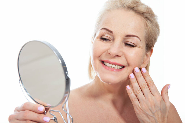 Happy middle aged woman model touching face skin looking in mirror. Smiling mature older lady enjoying healthy skin care, aging beauty, skincare treatment cosmetic products concept. - Photo, image
