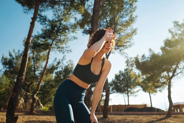 Young redhead woman wearing green sports bra and blue yoga pants standing on city park, after jogging catching her breath, wiping her sweat from her forehead. - 写真・画像