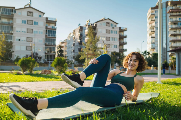 緑のスポーツブラジャーと青いヨガパンツを着た若い幸せな赤毛の女性は、屋外の都市公園のヨガマットに横たわっています。胸のポーズに膝の上に横たわる、 Apanasana運動を行う。アウトドアスポーツのコンセプト. - 写真・画像