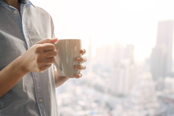 Κοντινό πλάνο χέρια γυναίκα κρατώντας λευκό φλιτζάνι καφέ ή τσάι από το παράθυρο με θολή θέα της πόλης υψηλό κτίριο στο παρασκήνιο. Αντιγραφή χώρου - Φωτογραφία, εικόνα